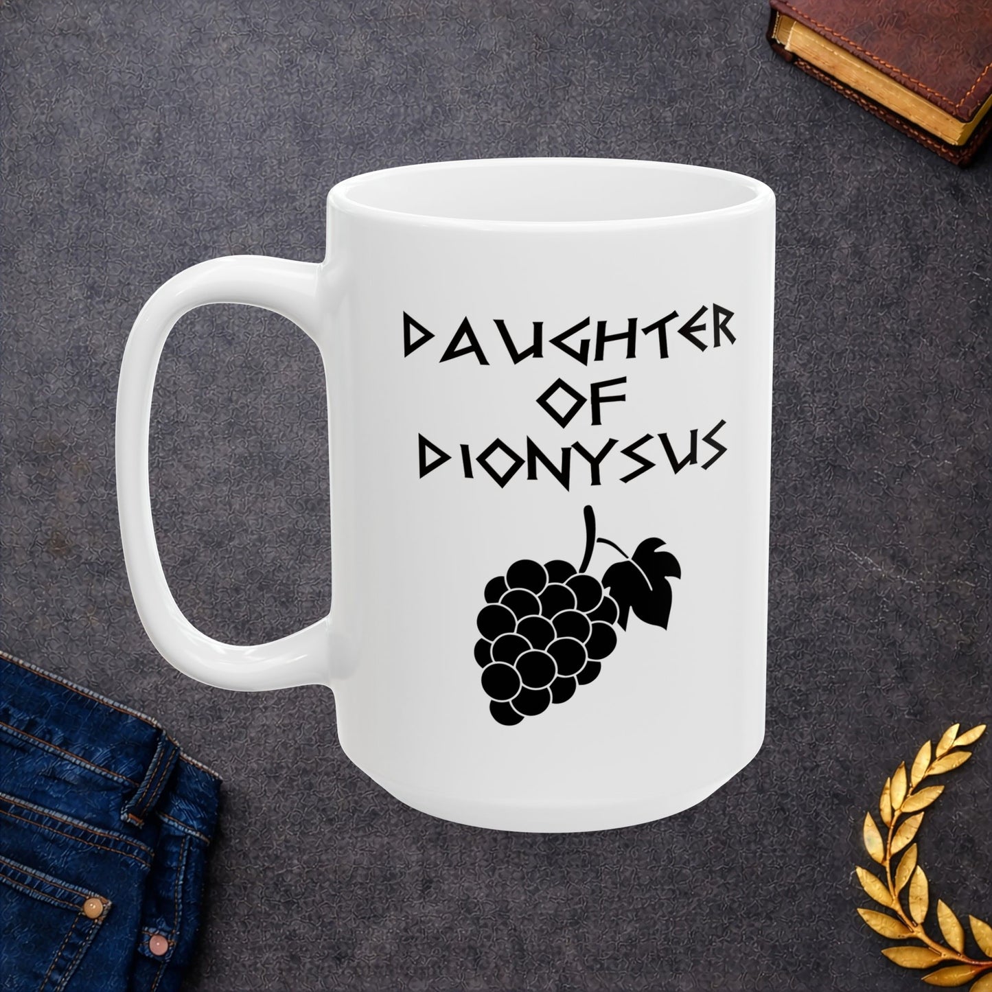 White ceramic mug featuring a minimalist, Greek-inspired graphic in black ink on a white background. The design reads 'DAUGHTER OF DIONYSUS' in a stylized, angular font, positioned above a clean, line-art graphic of a cluster of grapes hanging from a vine with leaves.