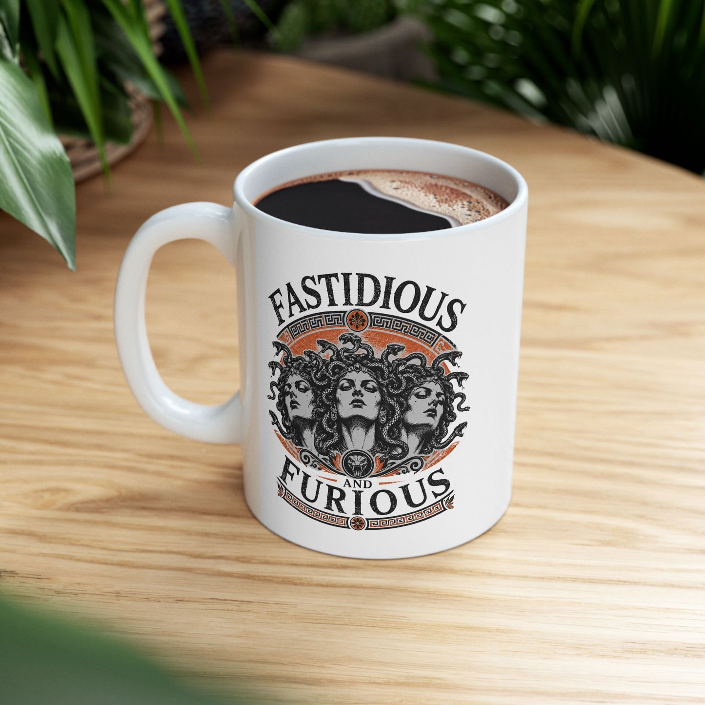 White ceramic mug with a Humorous design combining 'Fast and Furious' with Greek mythology on a white ceramic mug. Text reads 'FASTIDIOUS AND FURIOUS' in distressed lettering surrounding three faces of Medusa in black and white engraving style. Detailed serpents with individual scales form her hair. Greek key pattern borders in orange frame the composition with decorative sun motifs and a small snake head icon at center. Blends classical mythology with contemporary pop culture humor.