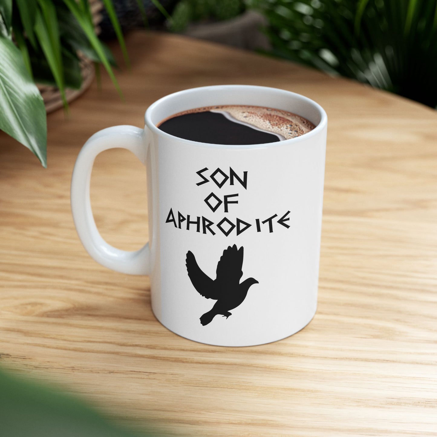 White ceramic mug featuring a minimalist, Greek-inspired graphic in black ink on a white background. The design reads 'SON OF APHRODITE' in a stylized, angular font, positioned above a clean, line-art graphic of a dove in flight.