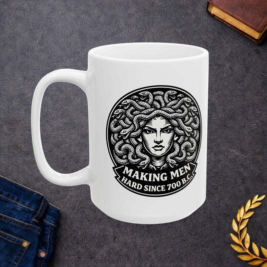 White ceramic mug with a bold black-and-white circular emblem featuring the head of Medusa with a mass of detailed, writhing snakes for hair. Below the illustration, a curved banner contains the humorous double-entendre "MAKING MEN HARD SINCE 700 B.C." written in a heavy, white serif font. The design uses a high-contrast, woodcut-inspired aesthetic with intricate line work against a dark background.