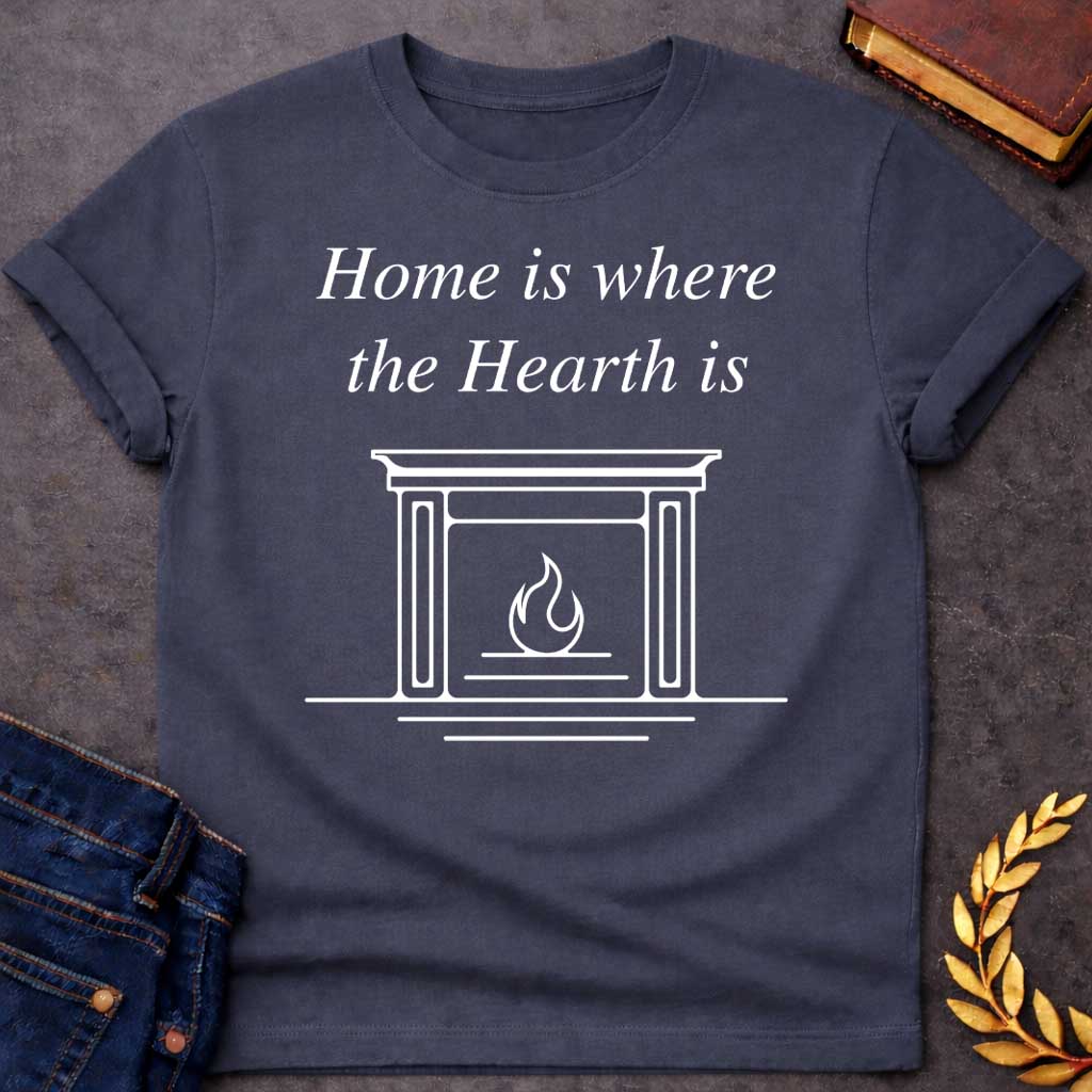Dark Heather T-shirt graphic in white featuring elegant italic serif text reading "Home is where the Hearth is" above a clean minimal line-art illustration of a fireplace mantel with a flame burning inside — a wordplay pun referencing Hestia, the Greek Goddess of the Hearth