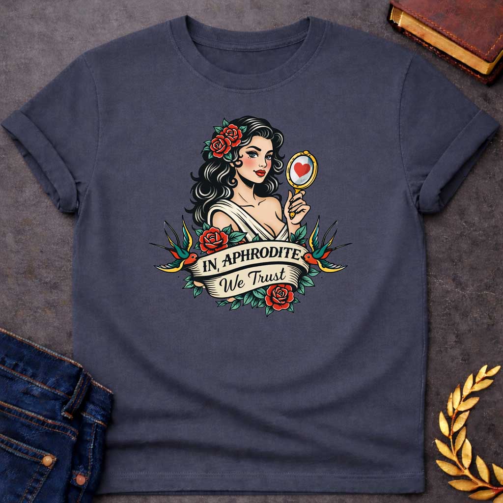 Dark Heather T-shirt graphic in a classic American traditional tattoo style featuring a pin-up illustration of Aphrodite, the Greek Goddess of Love, with dark wavy hair adorned with red roses, wearing a white draped gown, holding a gold hand mirror reflecting a red heart. She is flanked by two swooping swallows and surrounded by red roses and green leaves, with a scroll banner below reading "In Aphrodite We Trust" in bold and script lettering