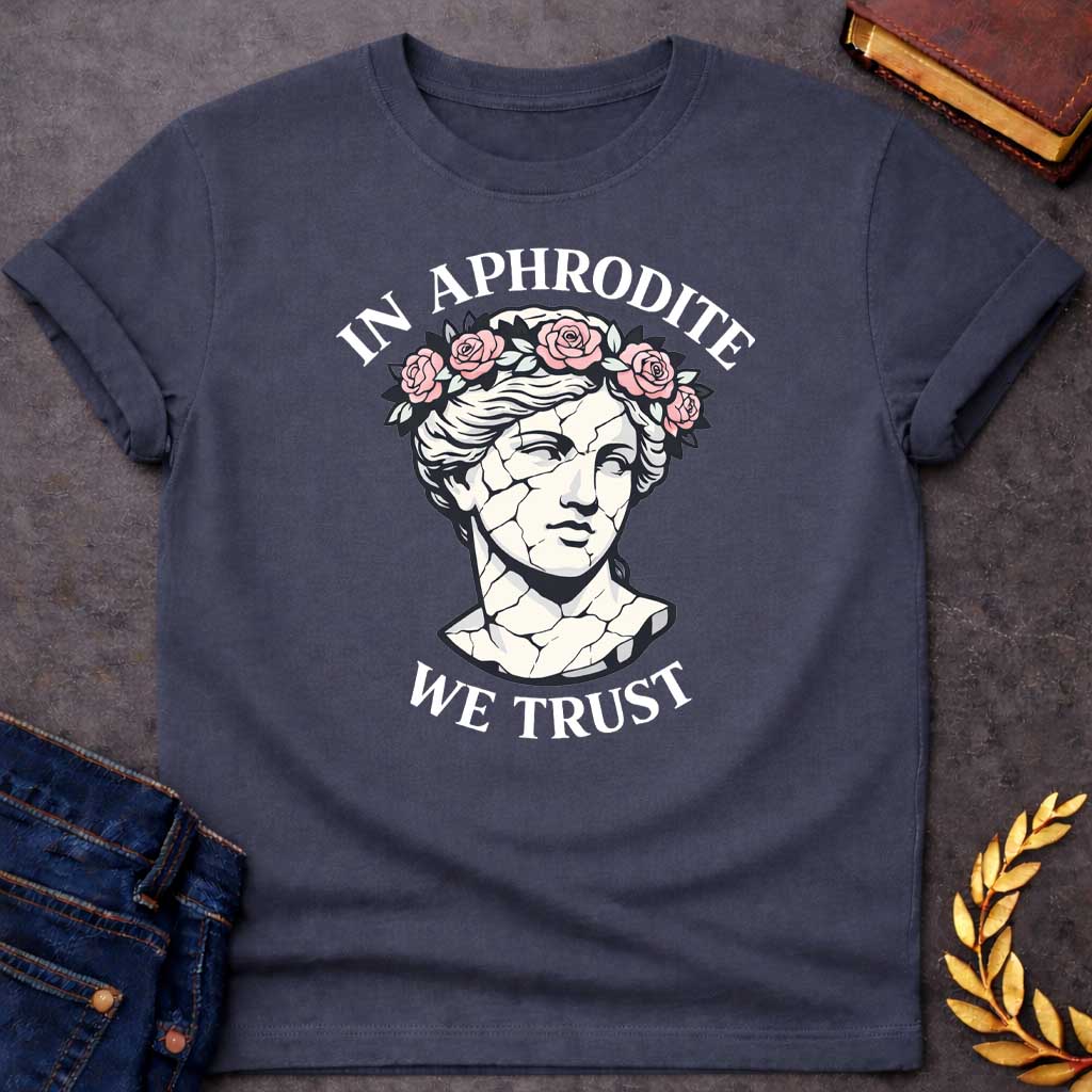 Dark Heather T-shirt graphic featuring a cracked marble bust illustration of Aphrodite, the Greek Goddess of Love, adorned with a floral crown of pink roses and green leaves, with bold arched text reading "In Aphrodite" curved above and "We Trust" curved below, in a modern aesthetic vaporwave-inspired style with white text and soft pink and white tones