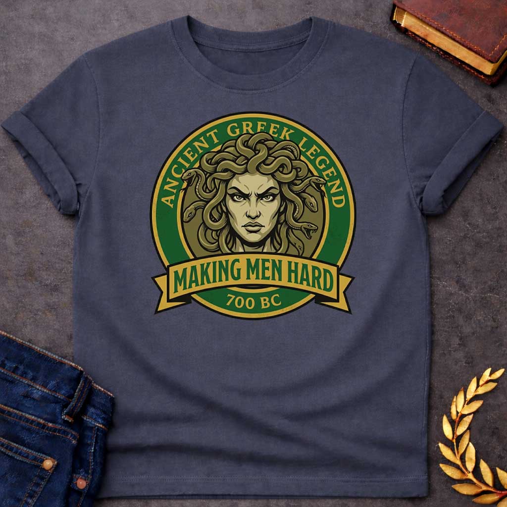 Dark Heather T-shirt graphic featuring a bold vintage badge design in forest green and gold. A fierce front-facing illustration of Medusa with coiling olive-toned snakes for hair and an intense glaring expression fills the circular center, with gold arched text reading "Ancient Greek Legend" around the upper border. A gold ribbon banner below the portrait reads "Making Men Hard" in green text, with "700 BC" below it — a witty double entendre referencing Medusa's mythological power to turn men to stone