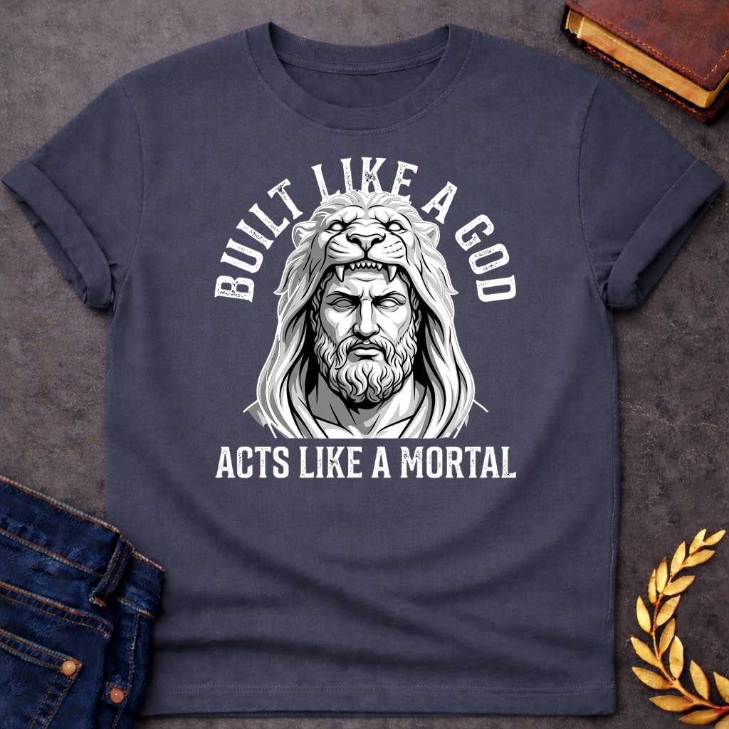 Dark Heather T-Shirt with a high-contrast, black-and-white graphic. The illustration depicts a stylized, statuesque man wearing the head and mane of a lion as a hood, reminiscent of Hercules. The text "BUILT LIKE A GOD" is arched over the top, and "ACTS LIKE A MORTAL" is printed in bold, straight text across the botto