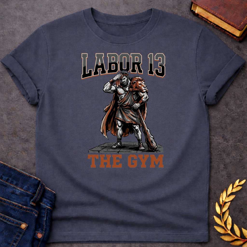 Dark Heather T-shirt featuring a sweat-drenched, muscular figure resembling Heracles, wearing a lion-skin cloak and holding a large wooden club. He stands on a stone platform while wiping his brow with a towel. The text "LABOR 13" is printed in large, bold, block letters across the top, and "THE GYM" is in a matching style at the bottom
