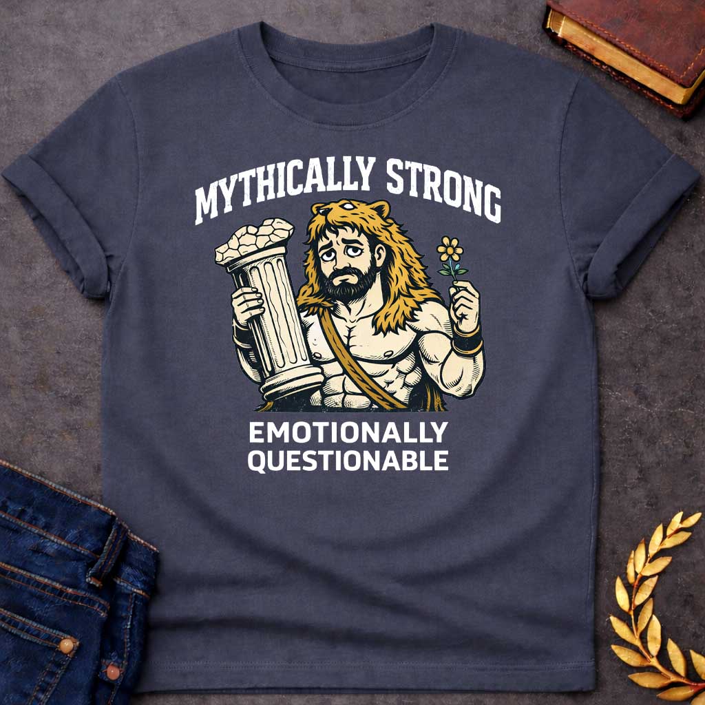 Dark Heather T-shirt featuring a humorous illustration of a muscular, bearded man wearing a lion-skin hood—reminiscent of Hercules—with a sad, weary expression. He holds a broken classical column in one hand and a small flower in the other. Arched over the top is the white text "MYTHICALLY STRONG," and below the illustration is the text "EMOTIONALLY QUESTIONABLE,"