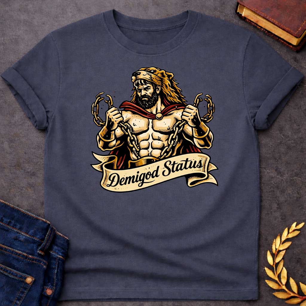 Dark Heather T-shirt featuring a muscular, bearded figure resembling Heracles, wearing a lion-skin hood and a red cape, as he heroically snaps thick golden chains with his hands. Below the illustration is a flowing, decorative banner with the text "Demigod Status" written in a stylish script font,