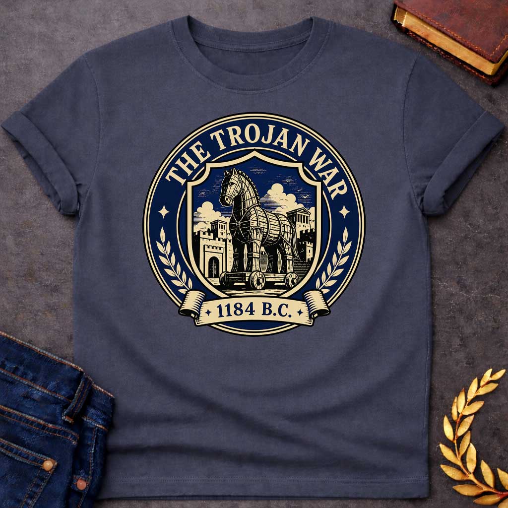 Dark Heather T-shirt with a graphic featuring a formal vintage seal design in navy and cream. A detailed engraving-style illustration of the Trojan Horse on wheels is centered within a shield, set against the walled city of Troy with Greek temple columns and dramatic clouds. Text arched around the outer circle reads "The Trojan War" with gold diamond stars, flanked by cream laurel branches, and a scroll banner below reads "1184 B.C." — referencing the legendary Trojan War from Greek mythology