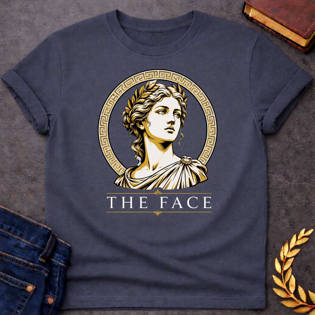 Dark Heather T-shirt with a classical Greek graphic featuring a gold and cream vector portrait of Helen of Troy in profile. She wears a golden laurel wreath and a traditional draped chiton, looking upward with a serene expression. The portrait is framed by a circular border with a gold Greek key (meander) pattern. Below the emblem, the text "THE FACE" is written in a clean, elegant white serif font between horizontal gold accent lines.