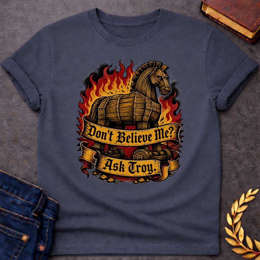 Dark Heather T-shirt with a bold illustration of the Trojan Horse engulfed in flames with red, orange, and yellow fire and black smoke. The wooden horse emerges from flames with detailed wood-grain texture. Two ornate gold banners with blackletter text read 'Don't Believe Me?' and 'Ask Troy.' Dark red background with rubble scattered below. References Cassandra's ignored warning about the Trojan Horse that led to Troy's destruction. Dramatic tattoo-inspired style with vintage engraving detail.