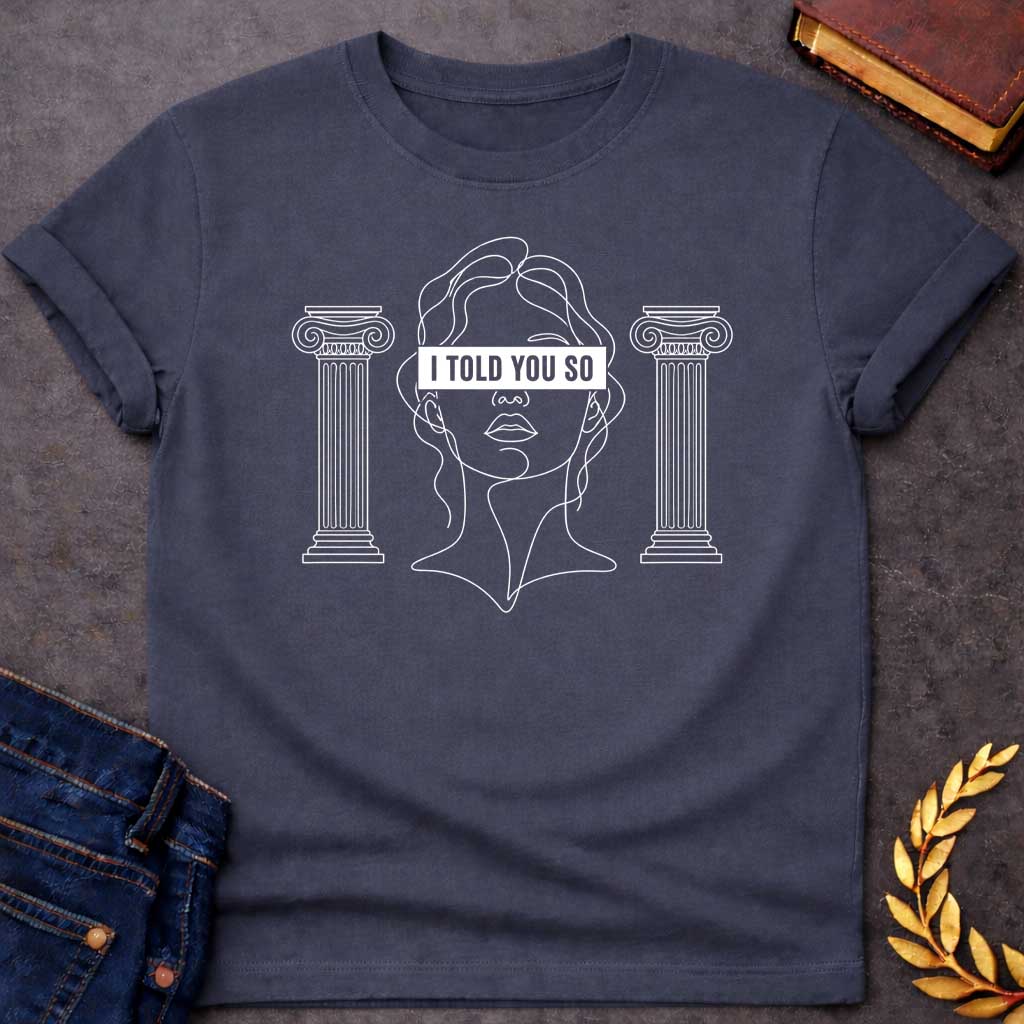 Dark Heather T-shirt with a minimalist line art design featuring a continuous white ink sketch of Cassandra or a Greek goddess. Her eyes are covered by a bold, solid black rectangular bar with the text "I TOLD YOU SO" in white sans-serif capital letters. The portrait is flanked on both sides by simple, symmetrical line-drawn Ionic Greek columns.