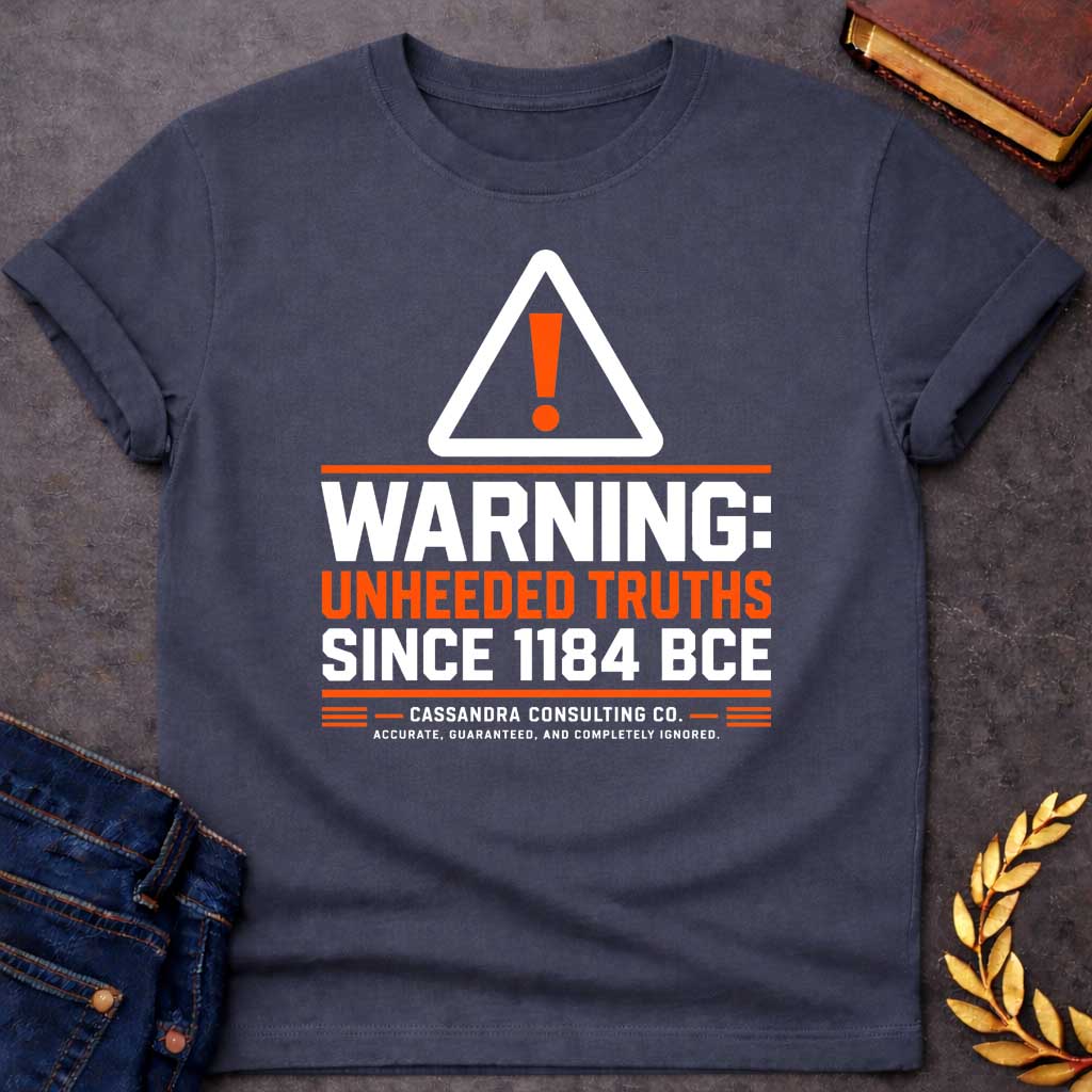 Dark Heather T-shirt with a corporate-style satirical design mimicking an industrial hazard sign. At the top is a triangular warning icon with an orange exclamation point. Bold block text reads "WARNING: UNHEEDED TRUTHS SINCE 1184 BCE" in alternating white and orange. Below, it features a faux company name: "CASSANDRA CONSULTING CO." followed by the tagline, "ACCURATE, GUARANTEED, AND COMPLETELY IGNORED."