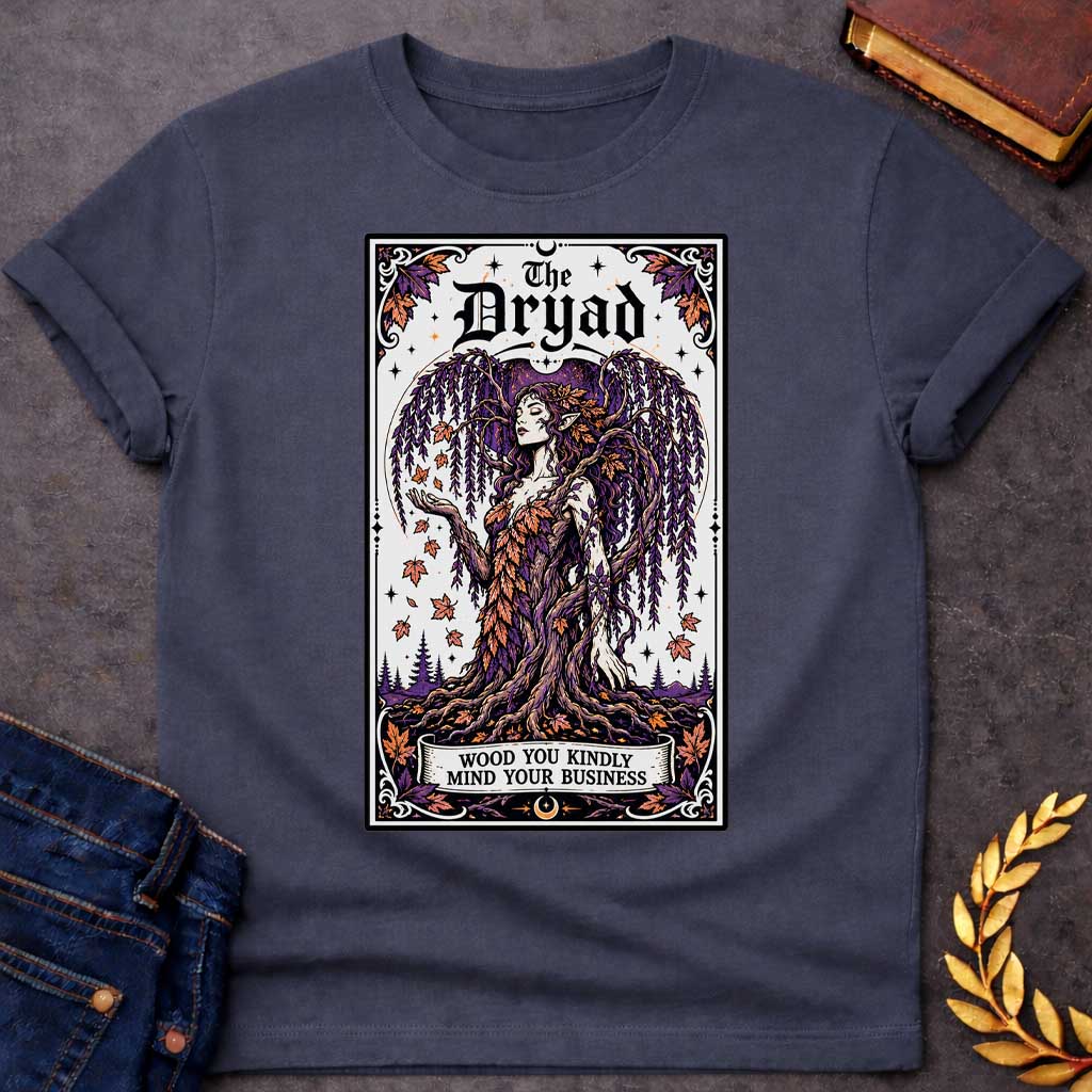 Dark Heather T-shirt with a mystical, tarot-card style design framed by an ornate border. It features a majestic weeping willow dryad whose gown is woven from purple and orange autumn leaves. At the top, bold gothic text reads "The Dryad." A weathered banner at the base of her rooted trunk features a clever, pun-based tagline that reads: "WOOD YOU KINDLY MIND YOUR BUSINESS" in a clean, black serif font