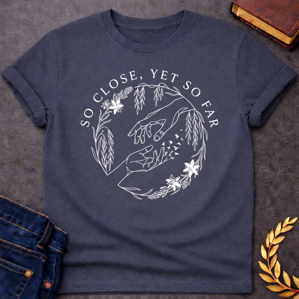 Dark Heather t-shirt in delicate fine-line style: two reaching hands almost touch, with one hand dissolving into a flock of small birds mid-fingertip, evoking Orpheus and Eurydice. Willow branches and small flowers form a circular wreath around the scene, with elegant serif text arcing above