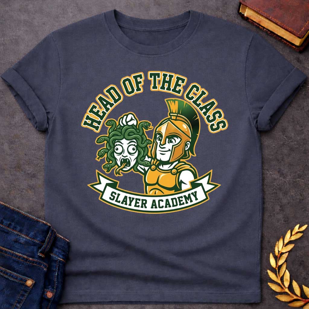 Dark Heather t-shirt in collegiate mascot style: a smirking gold-helmeted Greek warrior holds up Medusa's shocked, snarling severed head by her green snake hair. Bold green and gold text arcs above the design, with "Slayer Academy" on a white ribbon banner below. Playful varsity-logo aesthetic on a white background.