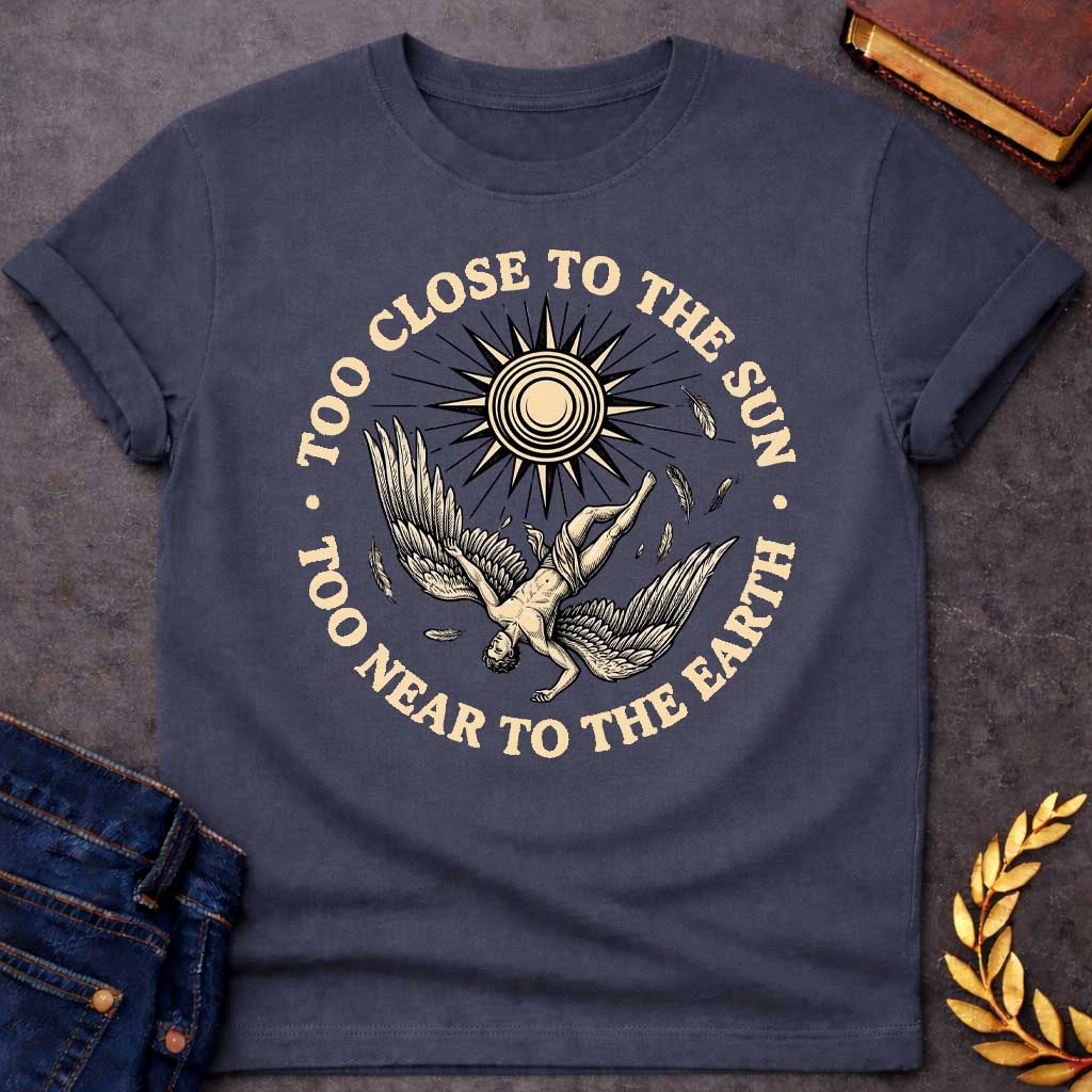 Dark Heather t-shirt with a vintage engraved-style illustration. A winged figure of Icarus plummets downward upside down as loose feathers drift through the air beneath a large, radiating sun emblem. Encircling the detailed central artwork, bold black serif text reads "TOO CLOSE TO THE SUN" along the top arch and "TOO NEAR TO THE EARTH" along the bottom curve.