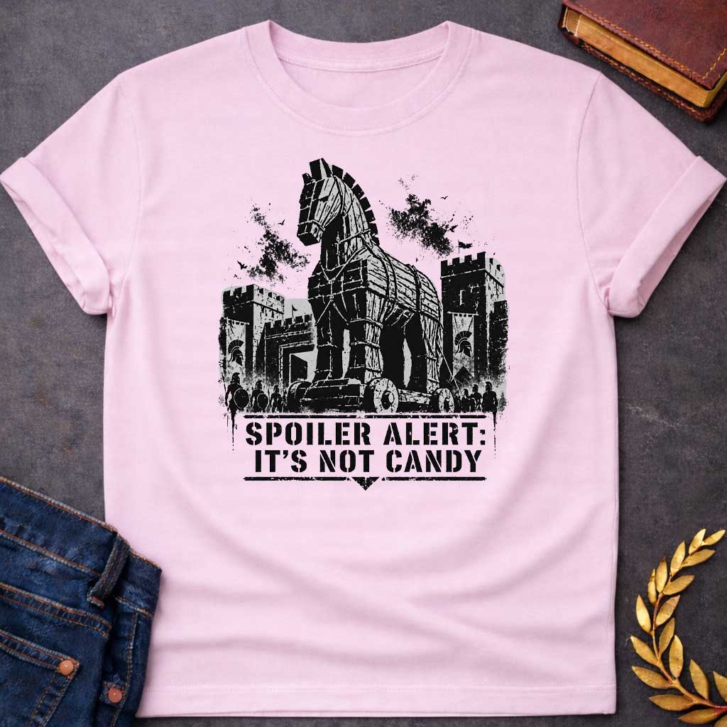 Light Pink T-shirt with a graphic in black featuring a gritty detailed ink splatter-style illustration of the Trojan Horse towering over the walls of Troy, flanked by Spartan warriors with shields and spears and castle battlements with birds circling overhead. Bold military stencil text in a distressed banner below reads "Spoiler Alert: It's Not Candy" — a humorous reference to the Greek legend of the Trojan War