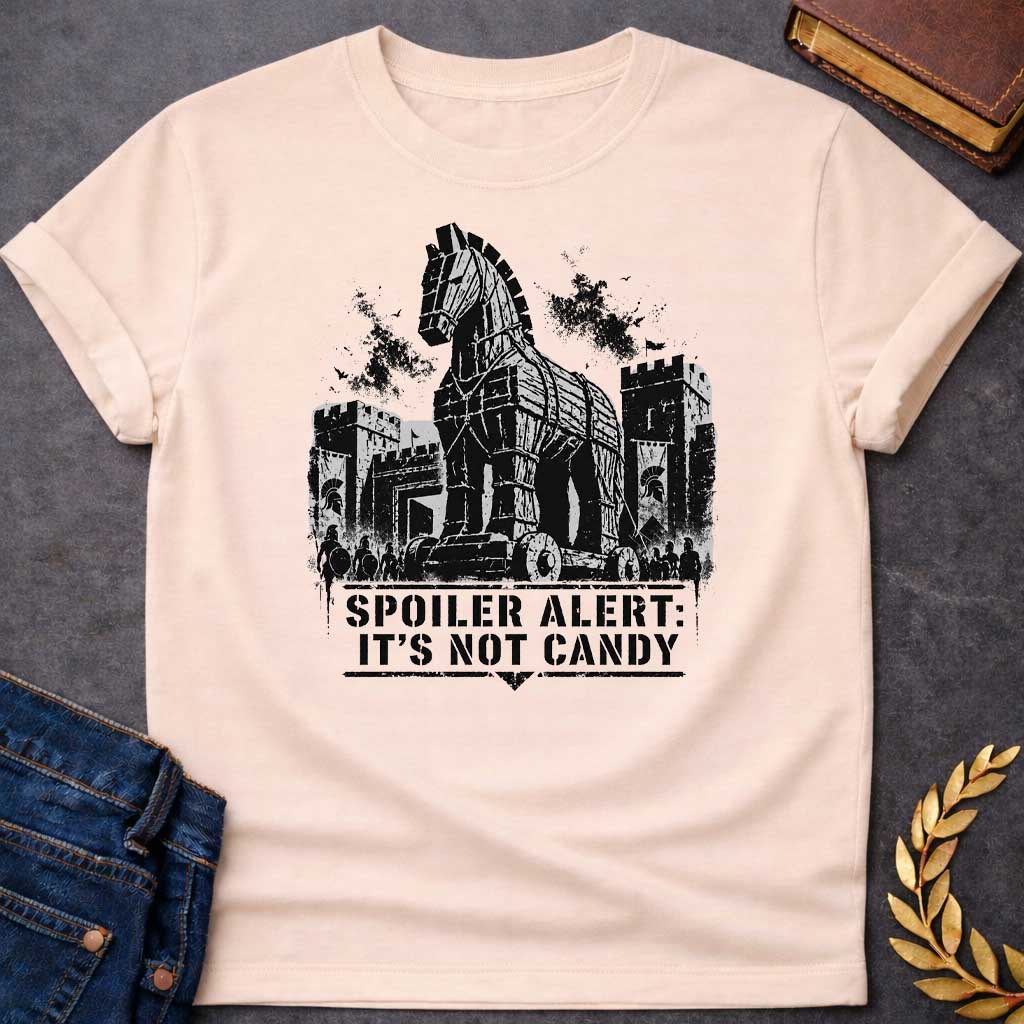Natural T-shirt with a graphic in black featuring a gritty detailed ink splatter-style illustration of the Trojan Horse towering over the walls of Troy, flanked by Spartan warriors with shields and spears and castle battlements with birds circling overhead. Bold military stencil text in a distressed banner below reads "Spoiler Alert: It's Not Candy" — a humorous reference to the Greek legend of the Trojan War