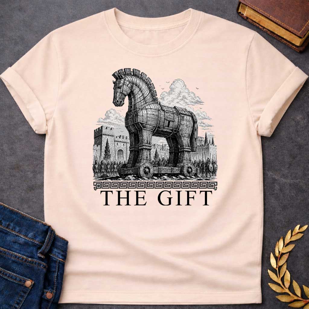 Natural T-shirt with a graphic in black featuring an exceptionally detailed fine-line engraving-style illustration of the Trojan Horse towering over the walls of Troy, surrounded by crowds of armored Greek soldiers with spears and shields, set against dramatic billowing clouds. A Greek meander border separates the scene from large bold classical serif text below reading "The Gift" — an ironic reference to the Greek legend of the Trojan War