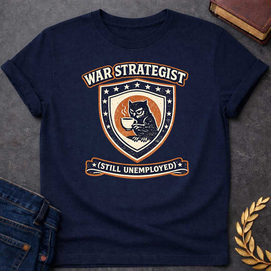 Navy T-shirt graphic featuring a vintage varsity-style crest in navy, orange, and cream. A grumpy owl holding a steaming coffee cup is centered on a shield surrounded by stars. An arched banner above reads "War Strategist" and a ribbon banner below reads "(Still Unemployed)," giving the design a humorous tone
