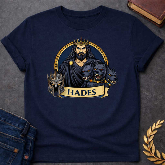 Navy T-shirt graphic featuring a vibrant illustrated portrait of Hades, the Greek God of the Underworld, with long dark wavy hair and a beard, piercing blue eyes, wearing a dark hooded cloak fastened with a gold brooch and a jeweled gold crown. He is flanked by the three snarling heads of Cerberus wearing a gold and gemstone collar, with a dark battle helmet displayed to the left. He is framed within a gold circular border against a navy background, with a scroll banner below reading "Hades," in a bold tatt