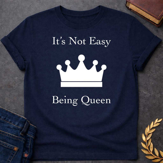 Navy T-shirt graphic in white on black featuring elegant serif text reading "It's Not Easy Being Queen" split above and below a simple white crown silhouette icon, in a humorous nod to Hera, the Greek Queen of the Gods