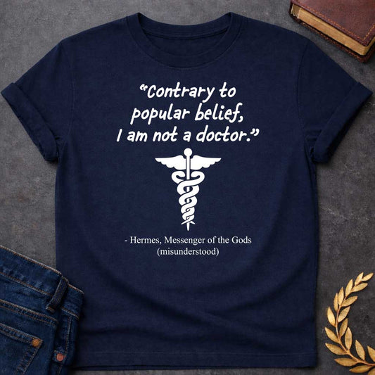 Navy T-shirt graphic in white text featuring a handwritten-style quote reading "Contrary to popular belief, I am not a doctor." above a white caduceus symbol, with the humorous attribution below in serif text reading "- Hermes, Messenger of the Gods (misunderstood)," playing on the common confusion between Hermes' caduceus and the medical symbol
