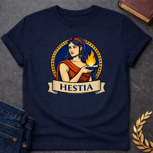 Navy T-shirt graphic featuring a vibrant illustrated portrait of Hestia, the Greek Goddess of the Hearth, with long dark blue wavy hair and a red headband, wearing gold drop earrings and a red draped robe, gently cradling a blue oil lamp with a glowing flame in her outstretched hands. She is framed within a gold circular border against a navy background, with a scroll banner below reading "Hestia," in a bold tattoo-inspired style with navy, gold, and red tones