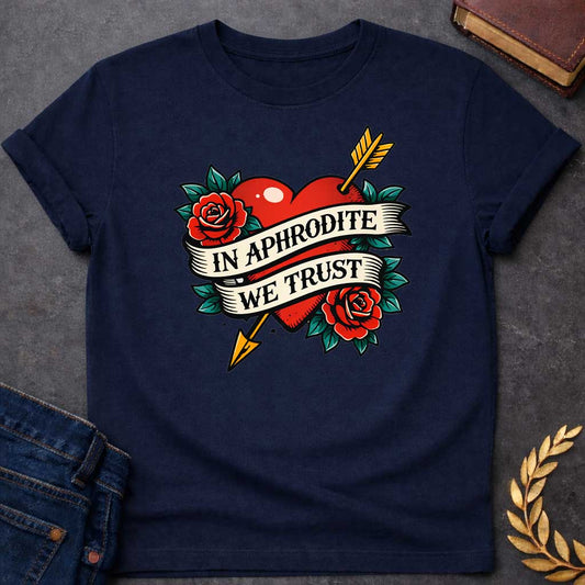 Navy T-shirt graphic in a classic American traditional tattoo style featuring a bold red heart pierced diagonally by a gold arrow, flanked by two red roses with teal green leaves, with a cream scroll banner wrapped around the heart reading "In Aphrodite We Trust" in black serif text — referencing Aphrodite, the Greek Goddess of Love, and Eros whose golden arrow inspired love