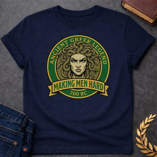 Navy T-shirt graphic featuring a bold vintage badge design in forest green and gold. A fierce front-facing illustration of Medusa with coiling olive-toned snakes for hair and an intense glaring expression fills the circular center, with gold arched text reading "Ancient Greek Legend" around the upper border. A gold ribbon banner below the portrait reads "Making Men Hard" in green text, with "700 BC" below it — a witty double entendre referencing Medusa's mythological power to turn men to stone