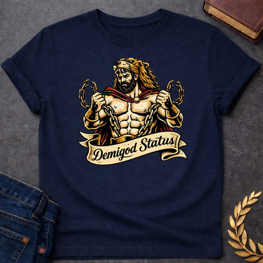 Navy T-shirt featuring a muscular, bearded figure resembling Heracles, wearing a lion-skin hood and a red cape, as he heroically snaps thick golden chains with his hands. Below the illustration is a flowing, decorative banner with the text "Demigod Status" written in a stylish script font,