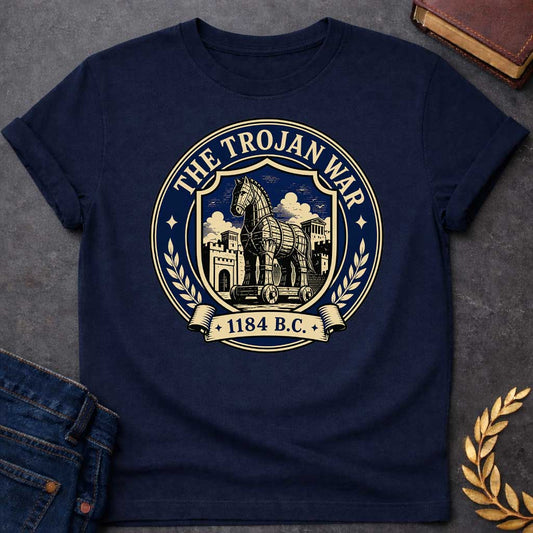 Navy T-shirt with a graphic featuring a formal vintage seal design in navy and cream. A detailed engraving-style illustration of the Trojan Horse on wheels is centered within a shield, set against the walled city of Troy with Greek temple columns and dramatic clouds. Text arched around the outer circle reads "The Trojan War" with gold diamond stars, flanked by cream laurel branches, and a scroll banner below reads "1184 B.C." — referencing the legendary Trojan War from Greek mythology
