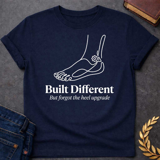 Navy T-Shirt with a minimalist line drawing of a human foot and ankle in dark gray profile, with a gear and a warning triangle icon near the heel. Below, bold serif text reads 'Built Different' and italicized text says 'But forgot the heel upgrade.' This design is a humorous, modern take on the myth of Achilles, playing on the slang 'built different' while joking that the hero's famous weakness was like a manufacturing defect or a forgotten software update