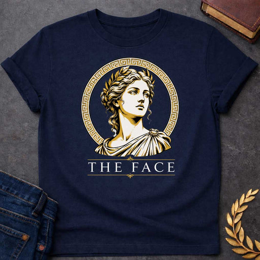 Navy T-shirt with a classical Greek graphic featuring a gold and cream vector portrait of Helen of Troy in profile. She wears a golden laurel wreath and a traditional draped chiton, looking upward with a serene expression. The portrait is framed by a circular border with a gold Greek key (meander) pattern. Below the emblem, the text "THE FACE" is written in a clean, elegant white serif font between horizontal gold accent lines.