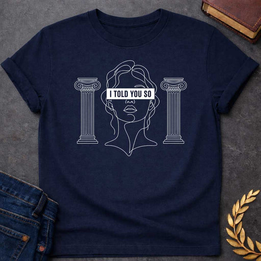 Navy T-shirt with a minimalist line art design featuring a continuous white ink sketch of Cassandra or a Greek goddess. Her eyes are covered by a bold, solid black rectangular bar with the text "I TOLD YOU SO" in white sans-serif capital letters. The portrait is flanked on both sides by simple, symmetrical line-drawn Ionic Greek columns.