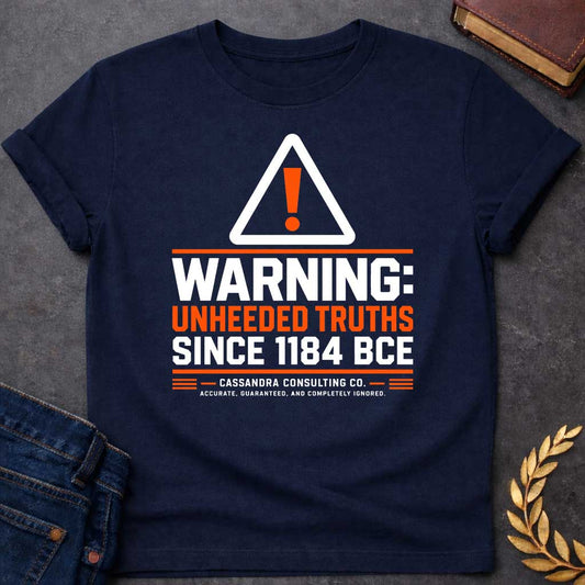 Navy T-shirt with a corporate-style satirical design mimicking an industrial hazard sign. At the top is a triangular warning icon with an orange exclamation point. Bold block text reads "WARNING: UNHEEDED TRUTHS SINCE 1184 BCE" in alternating white and orange. Below, it features a faux company name: "CASSANDRA CONSULTING CO." followed by the tagline, "ACCURATE, GUARANTEED, AND COMPLETELY IGNORED."