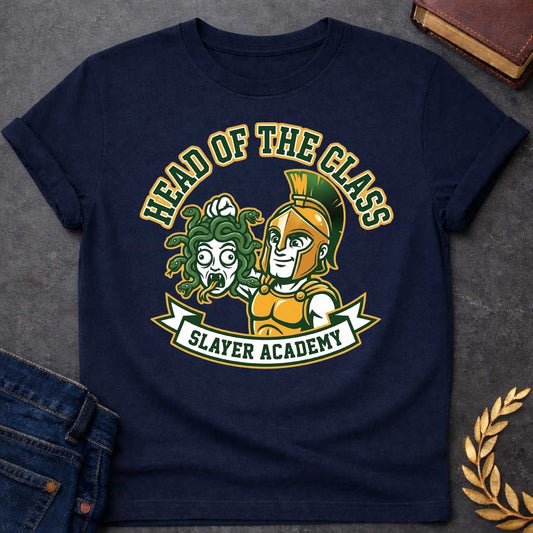 Navy t-shirt in collegiate mascot style: a smirking gold-helmeted Greek warrior holds up Medusa's shocked, snarling severed head by her green snake hair. Bold green and gold text arcs above the design, with "Slayer Academy" on a white ribbon banner below. Playful varsity-logo aesthetic on a white background.