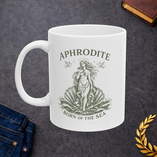 A white ceramic mug with a green illustration of Aphrodite on a shell and the text 'APHRODITE BORN OF THE SEA', shown on a dark textured background with jeans, a book, and a gold laurel leaf as styled props.