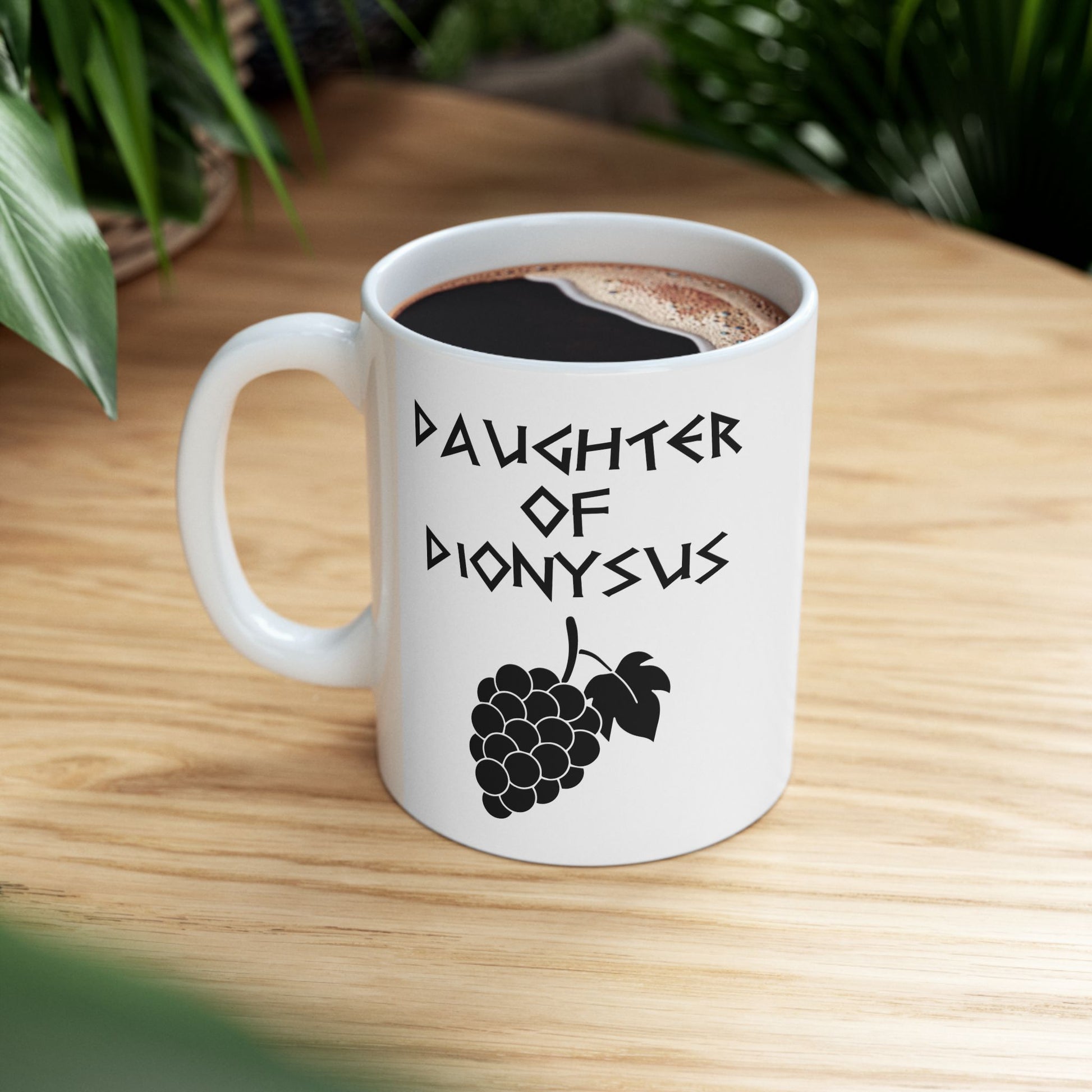 White ceramic mug featuring a minimalist, Greek-inspired graphic in black ink on a white background. The design reads 'DAUGHTER OF DIONYSUS' in a stylized, angular font, positioned above a clean, line-art graphic of a cluster of grapes hanging from a vine with leaves.
