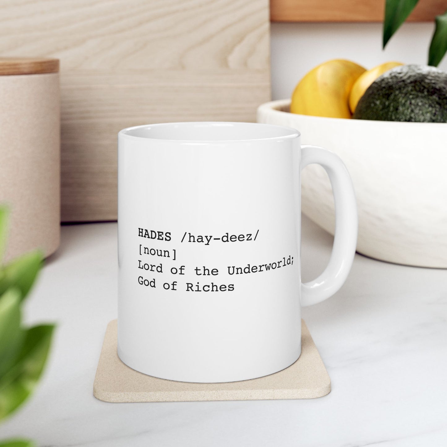 A minimalist graphic for a white ceramic mug featuring black typewriter-style text. The design is formatted like a dictionary entry for "HADES," including the phonetic pronunciation "/hay-deez/," the word class "[noun]," and the definition: "Lord of the Underworld; God of Riches.