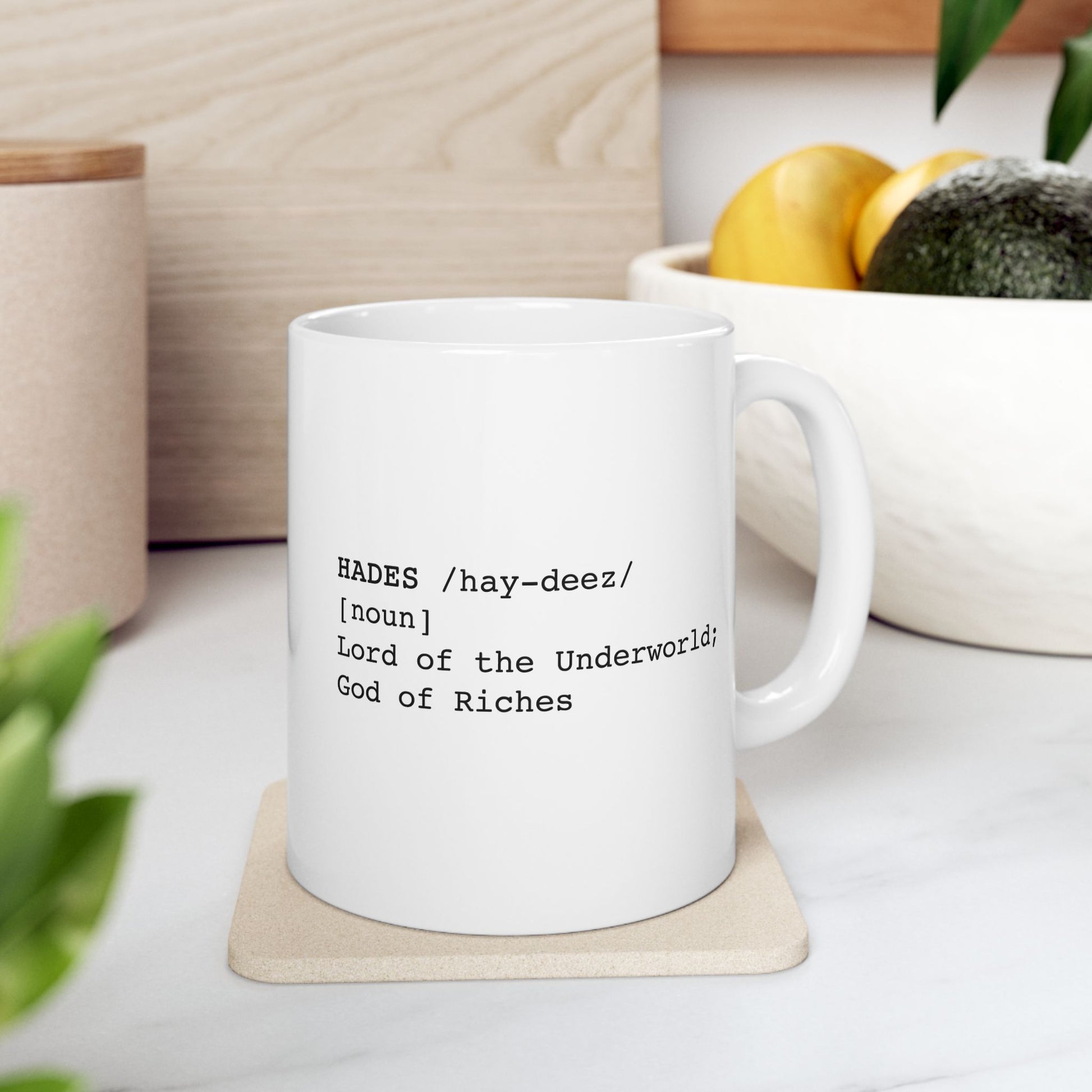 A minimalist graphic for a white ceramic mug featuring black typewriter-style text. The design is formatted like a dictionary entry for "HADES," including the phonetic pronunciation "/hay-deez/," the word class "[noun]," and the definition: "Lord of the Underworld; God of Riches.