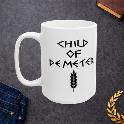 White ceramic mug featuring a minimalist, Greek-inspired graphic in black ink on a white background. The design reads 'CHILD OF DEMETER' in a stylized, angular font, positioned above a clean, line-art graphic of a bundle of harvested wheat.