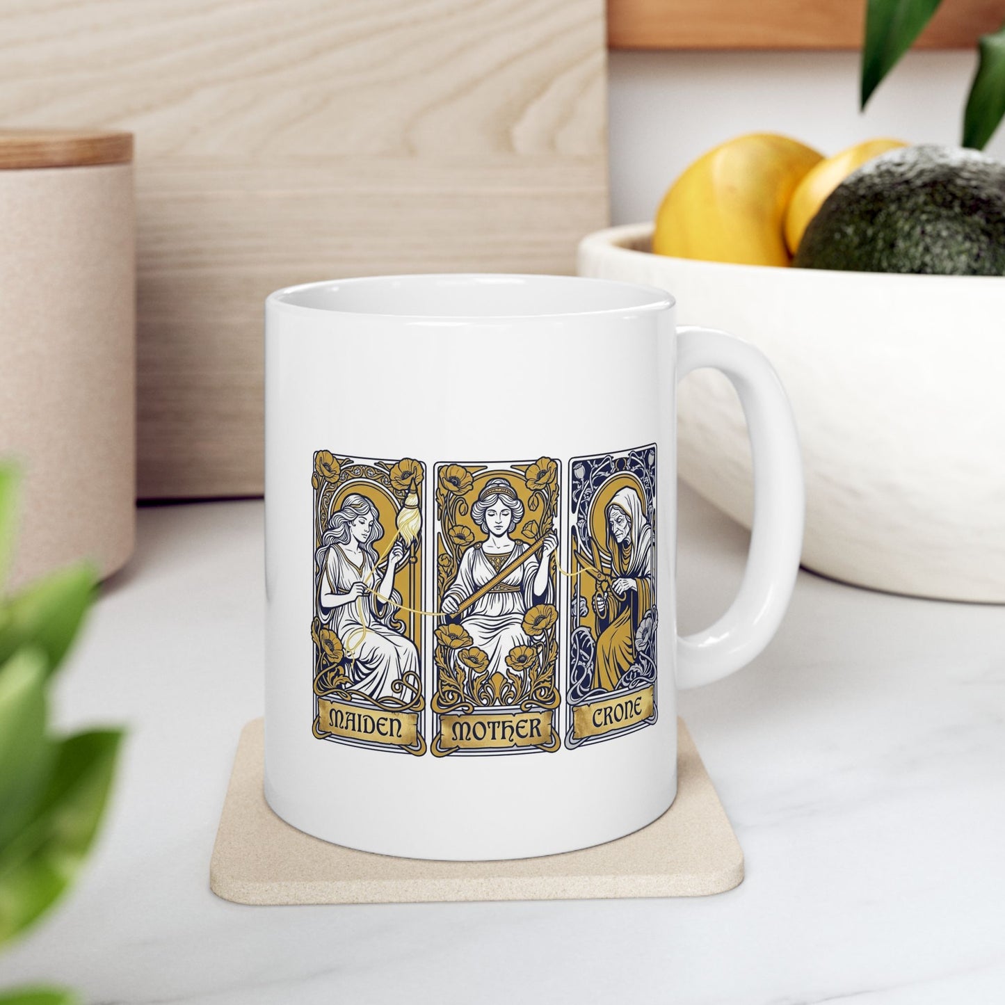 White ceramic mug with Three tarot-style cards depicting the triple goddess archetype: Maiden, Mother, and Crone. The maiden holds a bow surrounded by yellow poppies, the mother wears a crown with a sword, and the crone wears hooded robes with a staff. Each rendered in Art Nouveau style with ornate decorative borders, gold halos, and floral flourishes. Labels appear at card bases. Celebrates the feminine divine in all life stages and cyclical transformation.