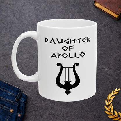 White ceramic mug featuring a minimalist, Greek-inspired graphic in black ink on a white background. The design reads 'DAUGHTER OF APOLLO' in a stylized, angular font, positioned above a clean, line-art graphic of a classical lyre.