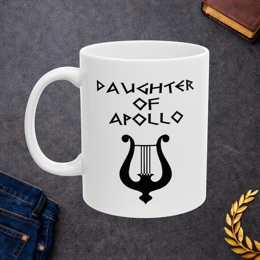 White ceramic mug featuring a minimalist, Greek-inspired graphic in black ink on a white background. The design reads 'DAUGHTER OF APOLLO' in a stylized, angular font, positioned above a clean, line-art graphic of a classical lyre.