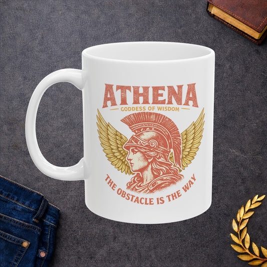 A white ceramic coffee mug with a red and gold graphic of Athena, the goddess of wisdom, with the text 'ATHENA GODDESS OF WISDOM' and 'THE OBSTACLE IS THE WAY'. The mug is displayed on a dark gray textured surface with styled props including blue jeans, a gold laurel wreath, and a leather journal.