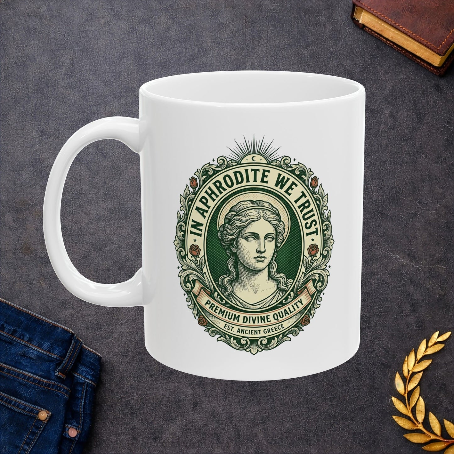 White ceramic mug with a vintage, currency-inspired emblem featuring a detailed bust of the goddess Aphrodite. The design is framed by an ornate oval border with roses and scrolls. The text "IN APHRODITE WE TRUST" arches above, while banners below read "PREMIUM DIVINE QUALITY" and "EST. ANCIENT GREECE." The artwork uses a classic green and cream color palette with a textured, engraved aesthetic.