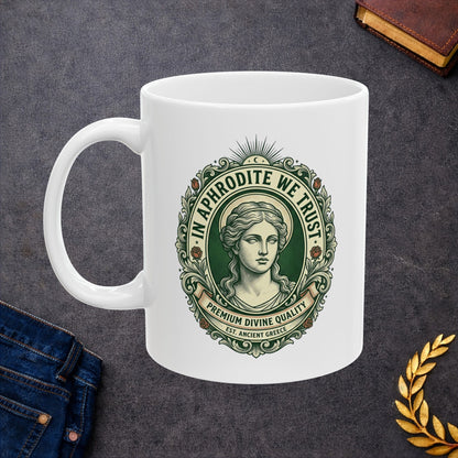 White ceramic mug with a vintage, currency-inspired emblem featuring a detailed bust of the goddess Aphrodite. The design is framed by an ornate oval border with roses and scrolls. The text "IN APHRODITE WE TRUST" arches above, while banners below read "PREMIUM DIVINE QUALITY" and "EST. ANCIENT GREECE." The artwork uses a classic green and cream color palette with a textured, engraved aesthetic.