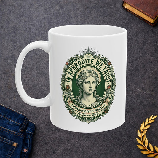 White ceramic mug with a vintage, currency-inspired emblem featuring a detailed bust of the goddess Aphrodite. The design is framed by an ornate oval border with roses and scrolls. The text "IN APHRODITE WE TRUST" arches above, while banners below read "PREMIUM DIVINE QUALITY" and "EST. ANCIENT GREECE." The artwork uses a classic green and cream color palette with a textured, engraved aesthetic.